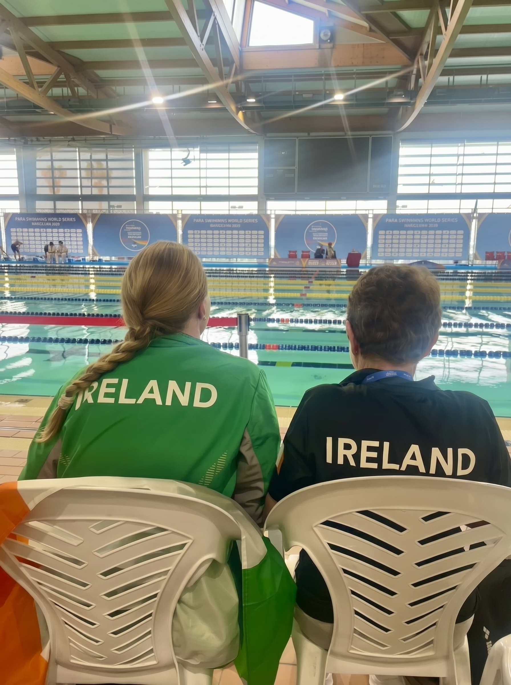 Two members of the Ireland team in tricolour jackets watching a swim session from poolside, with the tricolour draped at the lane edge