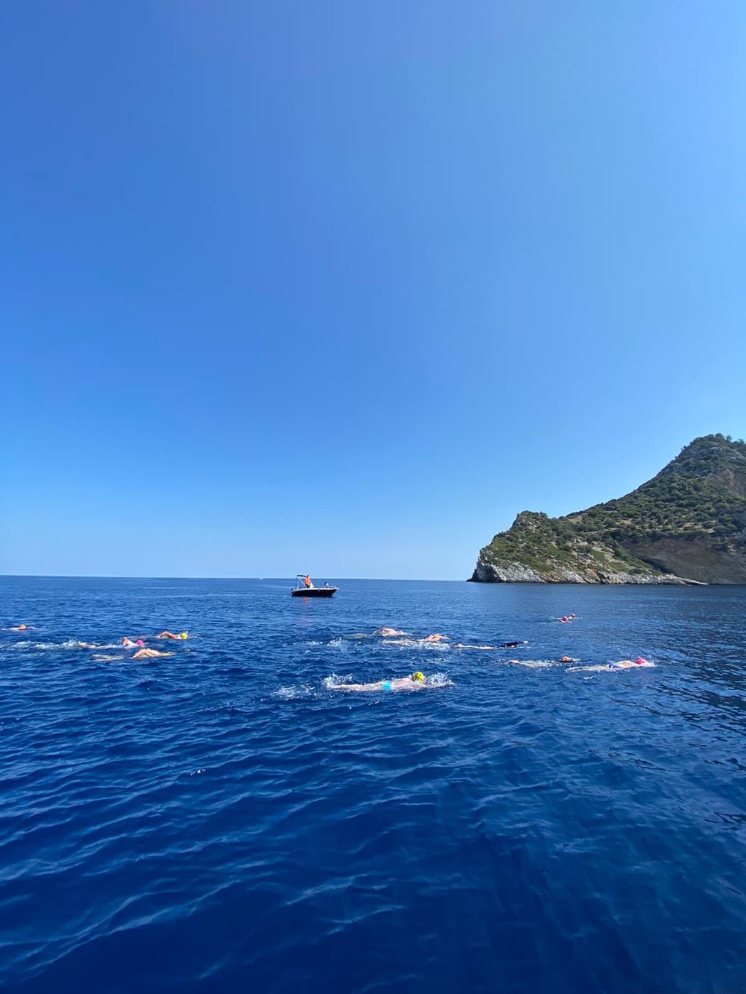 Open-water swimmers training in the sea alongside a safety boat, heading toward a headland
